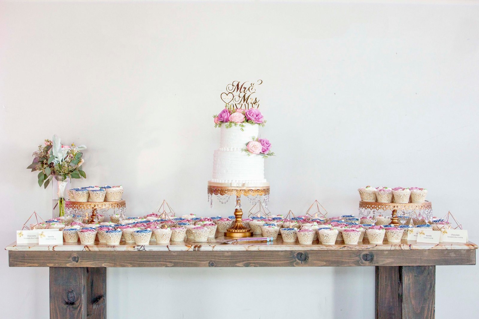 white wedding cake with pink flowers surrounded by cupcakes on a wood table