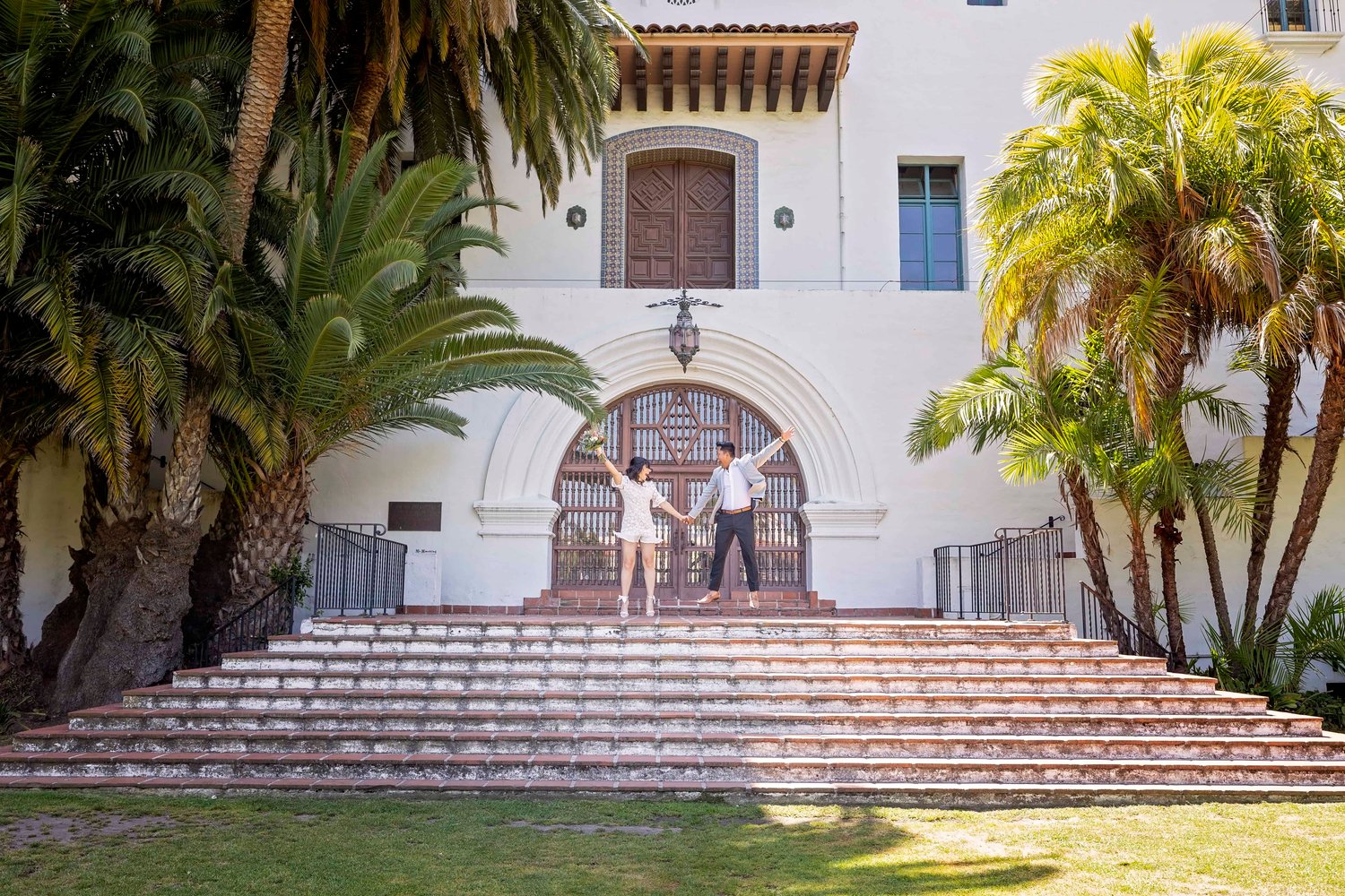 bride and groom jumping on the steps in front of an arched doorway at the Santa Barbara Courthouse