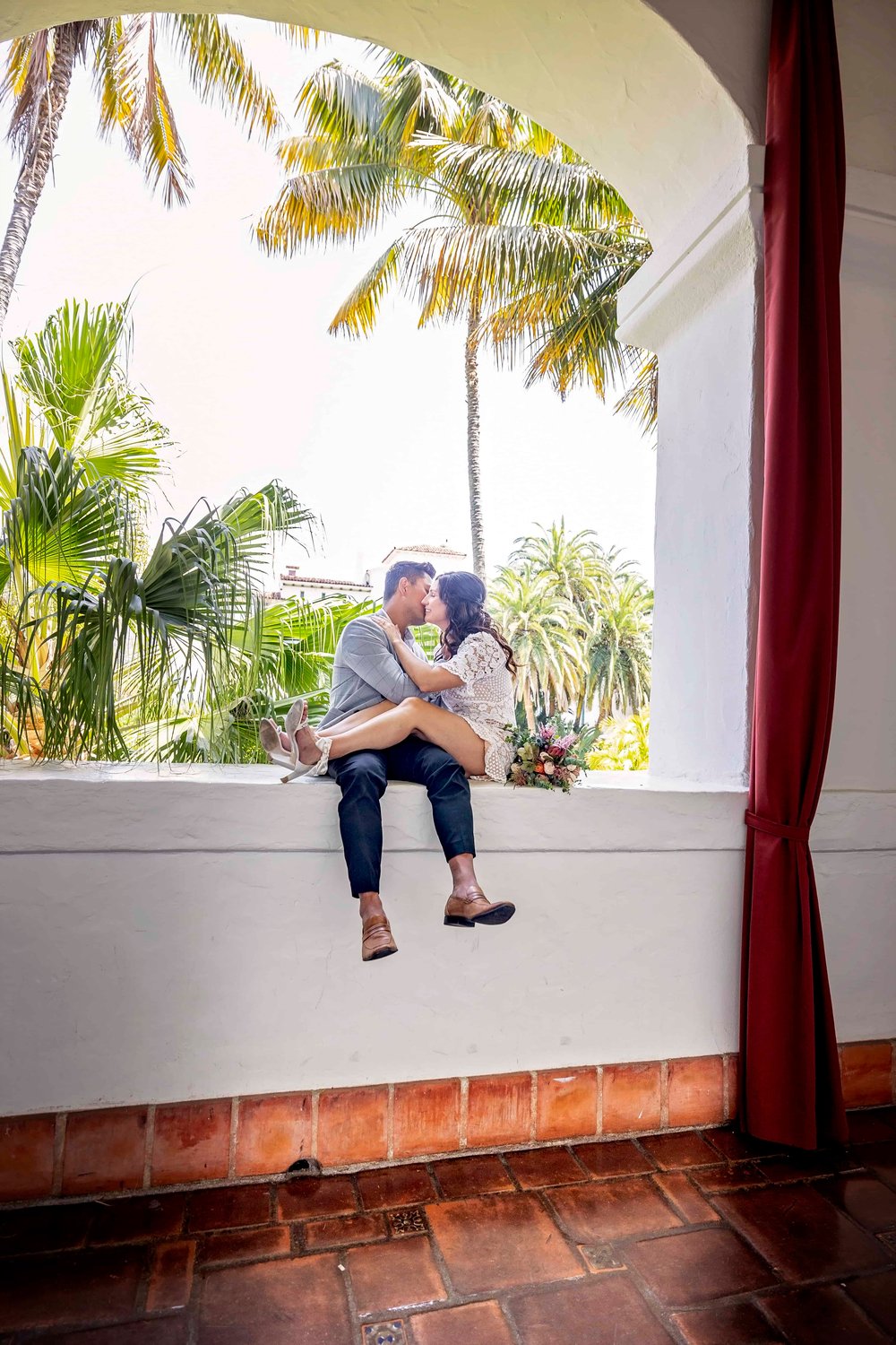 bride and groom kissing while sitting on a ledge with palm trees behind them