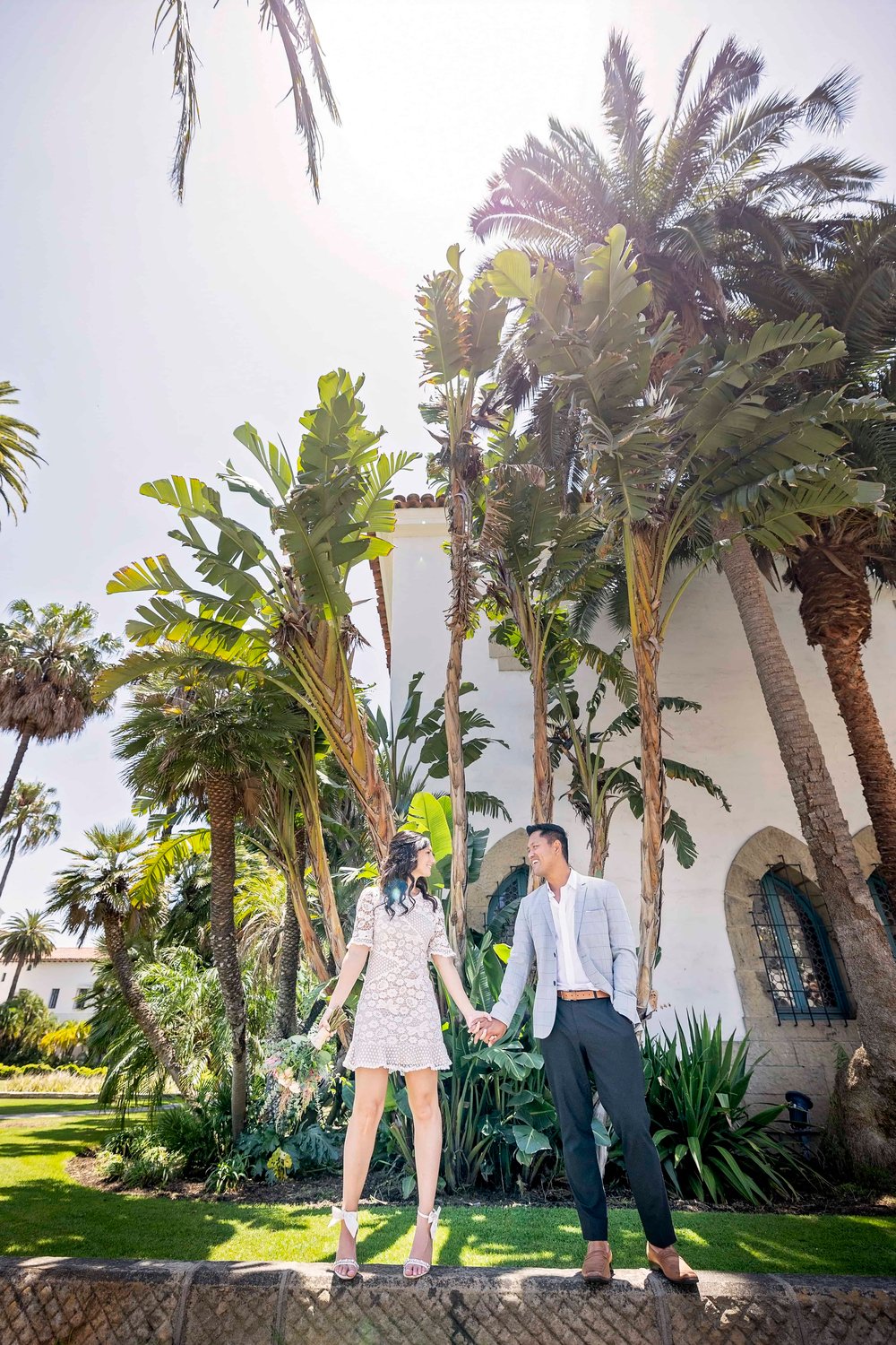 bride and groom standing on a short wall with palm trees and the Santa Barbara Courthouse behind them
