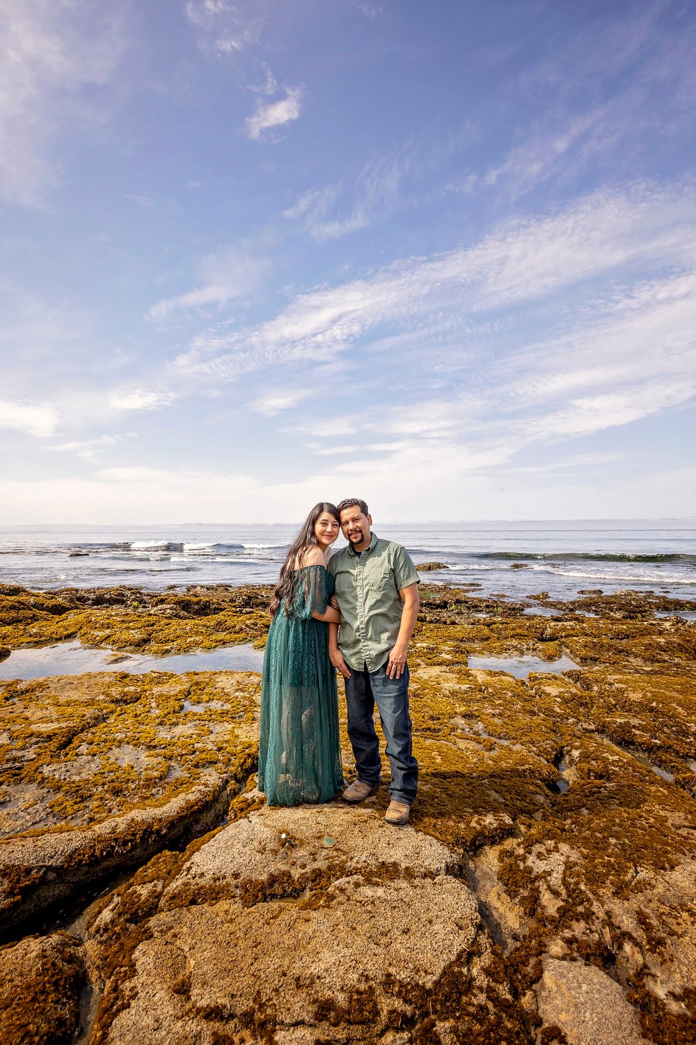 couple standing on rocks at the beach while looking at the camera during an engagement photoshoot