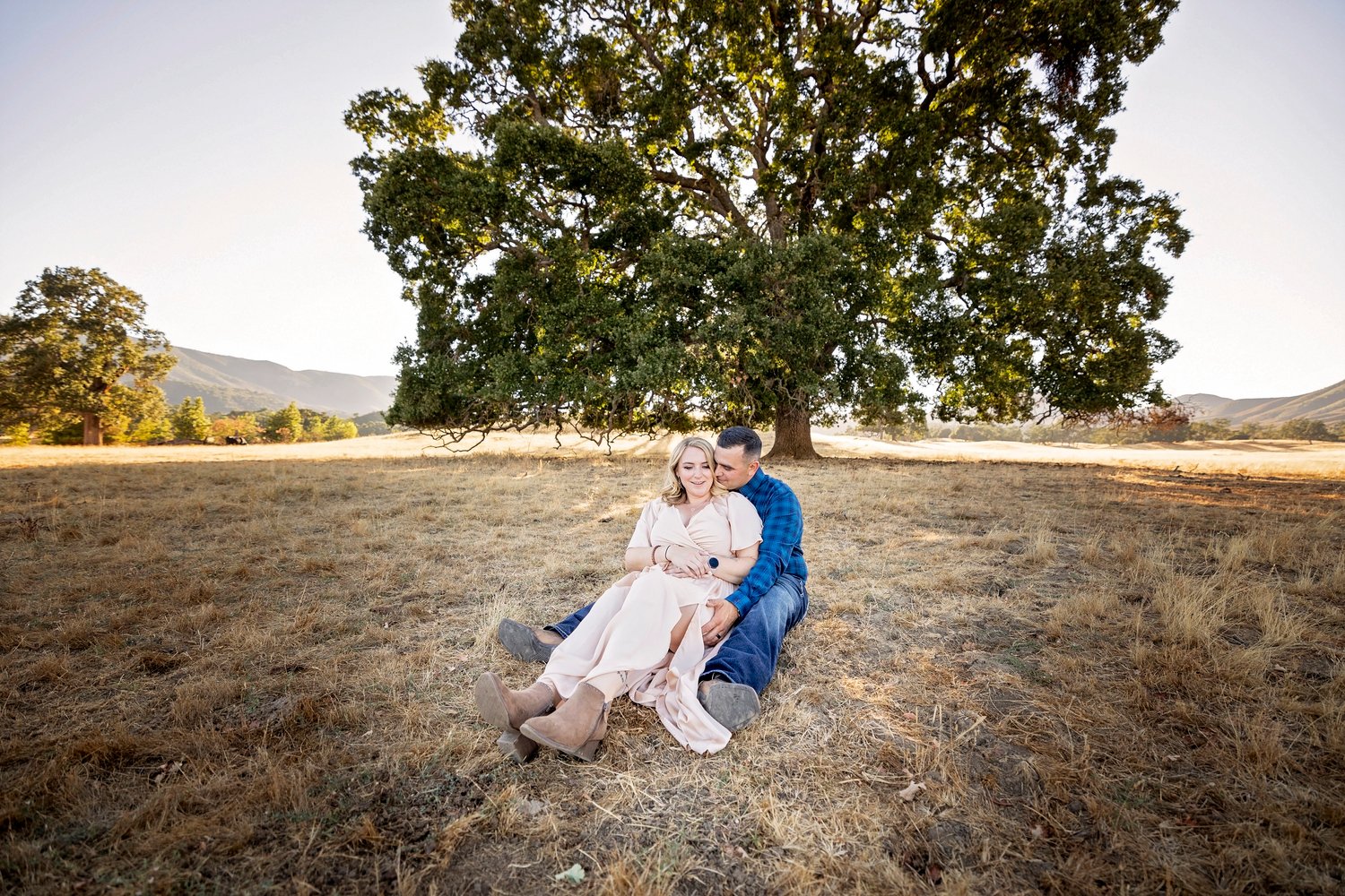 couple sitting on the ground and cuddling in a field in front of an oak tree