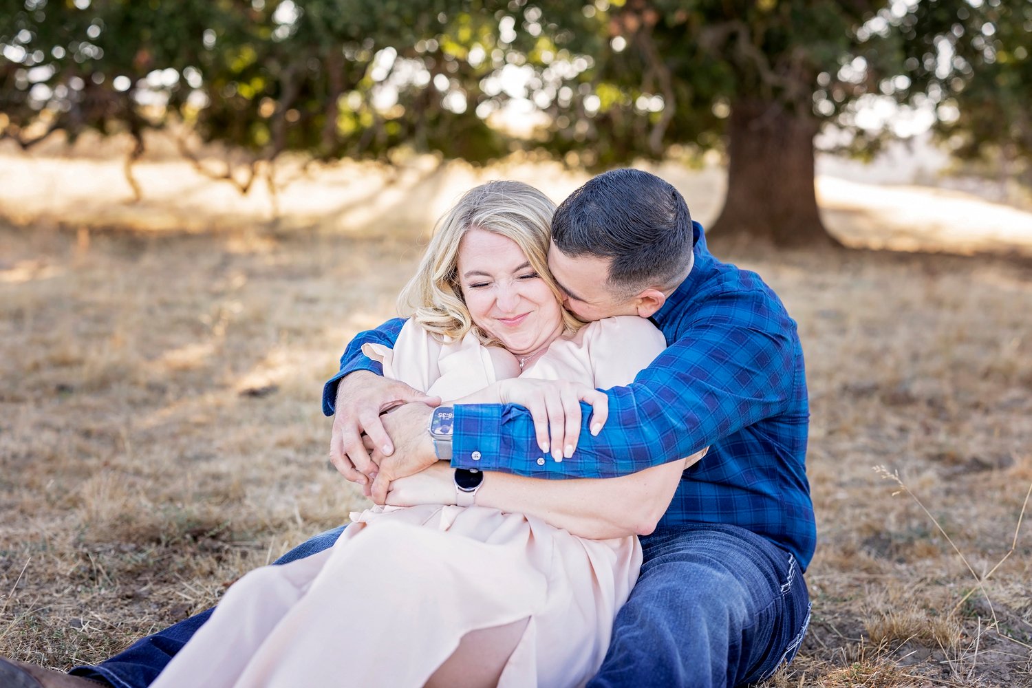 couple sitting on the ground and cuddling in a field in front of an oak tree