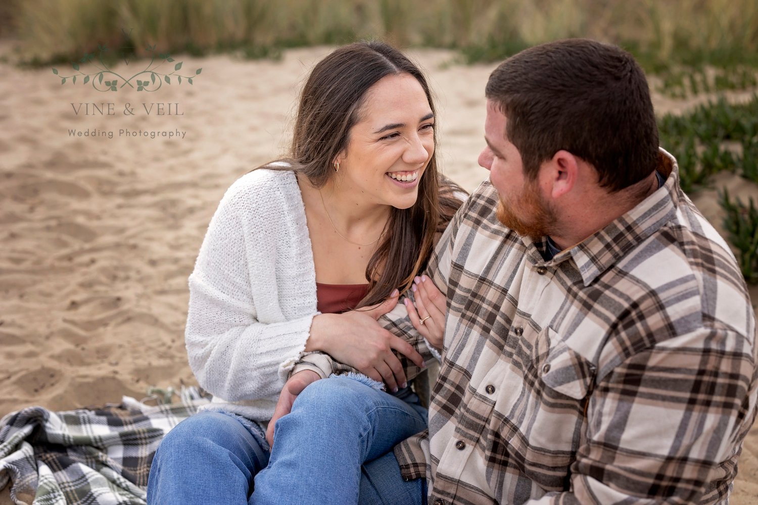 woman smiling at her boyfriend while they sit together in the sand at the beach