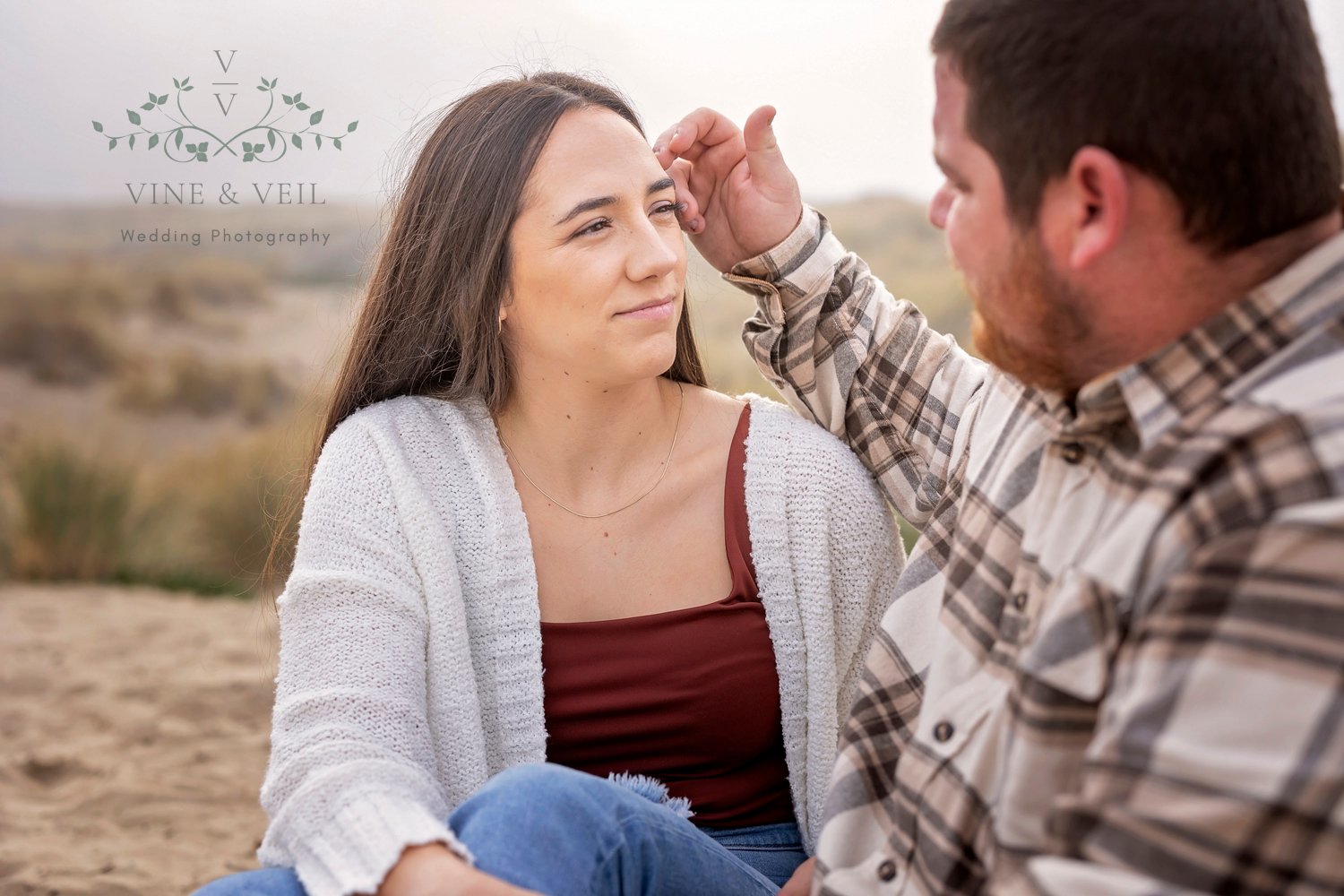 man moving the hair out of his girlfriend's face while they sit together on the sand