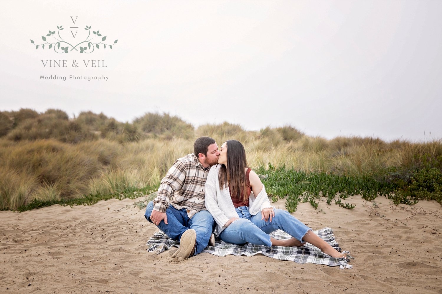 couple kissing while sitting on a blanket in the sand with beach grass behind them
