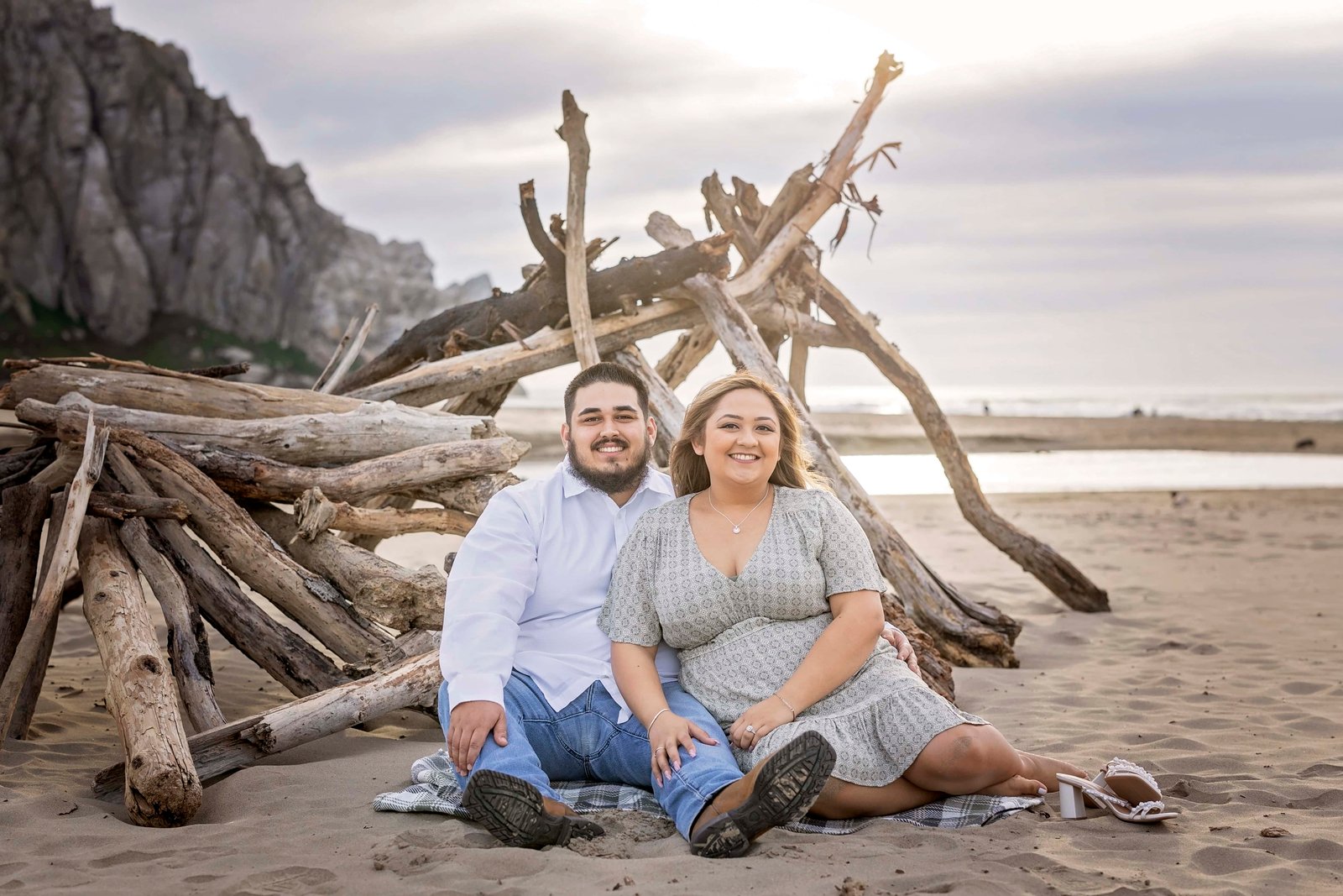 couple smiling at the camera while sitting in the sand in front of some driftwood