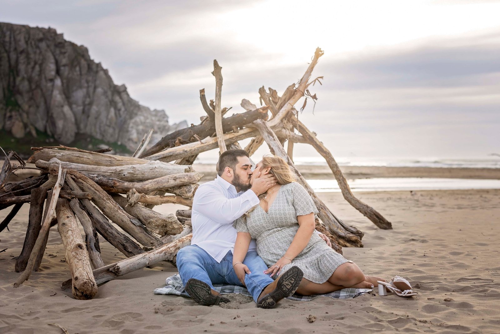 couple kissing while sitting in the sand in front of some driftwood